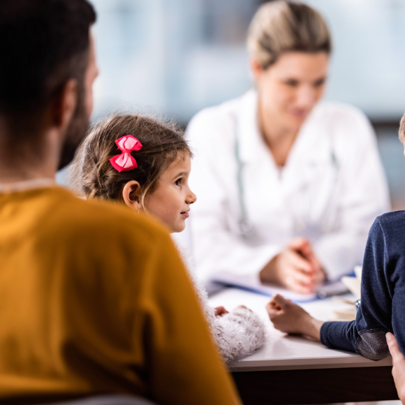 Ein Mädchen sitzt auf dem Schoß ihres Vaters, welche von hinten fotografiert wird, während sie zur Seite schaut, wo ihr Bruder sitzt. Im Hintergrund sieht man eine Ärztin, welche sich mit der Familie unterhält.