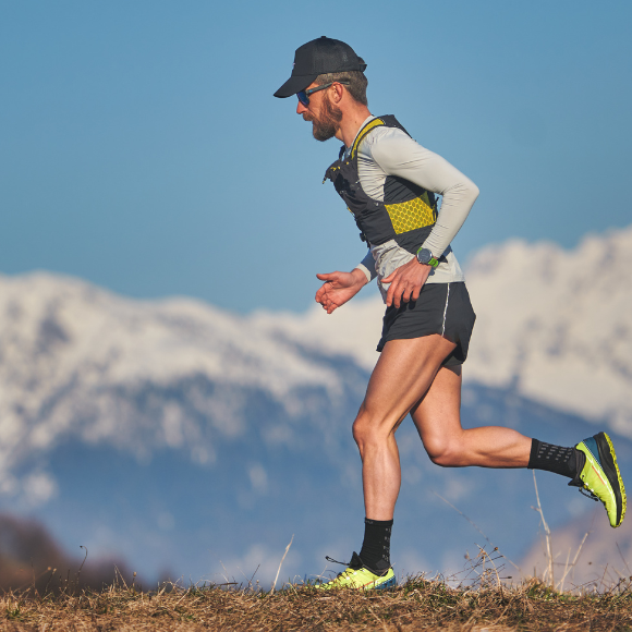 Ein Mann mit Sonnenbrille und kurzer Hose joggt vor einem Panorama auf Bergen mit einer Schneespitze.