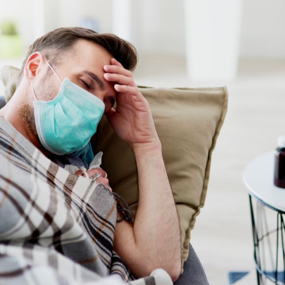 Ein Mann liegt mit Maske und einer Decke zugedeckt auf einem Sofa und hält sich den Kopf.