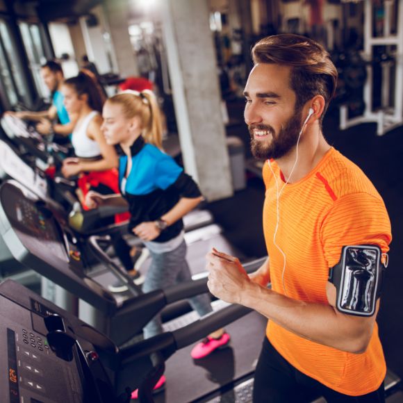 Ein Mann in orangenem Shirt läuft auf einem Laufband und lacht. Neben ihm sind noch weitere Personen auf Laufbändern zu sehen.