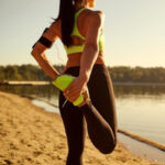 Eine Frau steht in Sport-Outfit vor einem See und dehnt sich